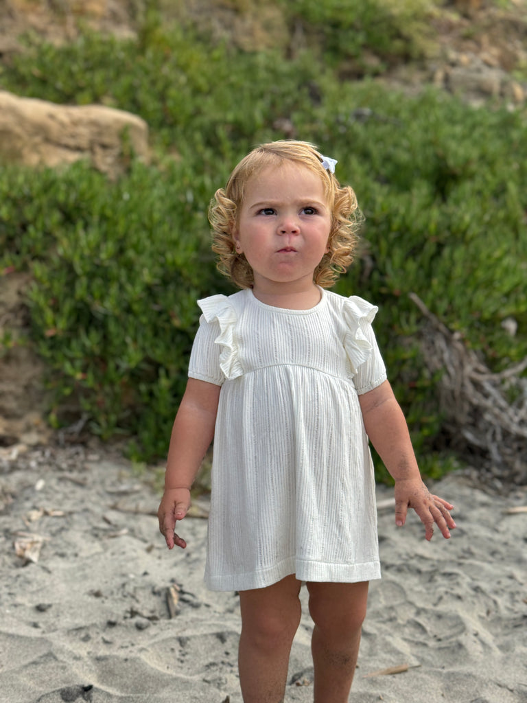 happy toddler standing on beach on sunny day wearing white dress with frill over shoulders and short sleeves line pattern fabric