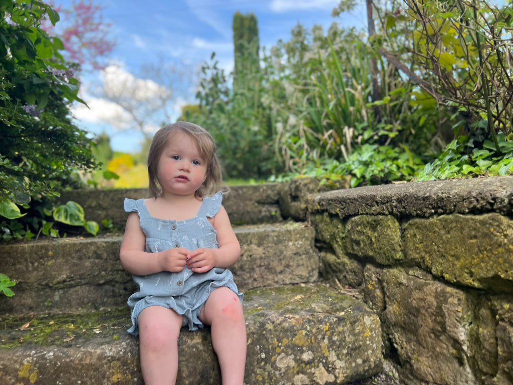 toddler sitting on old stone steps in garden wearing gentle blue gauze shortie overalls with white all over seahorse print. buttons on front and frill detail round front and legs