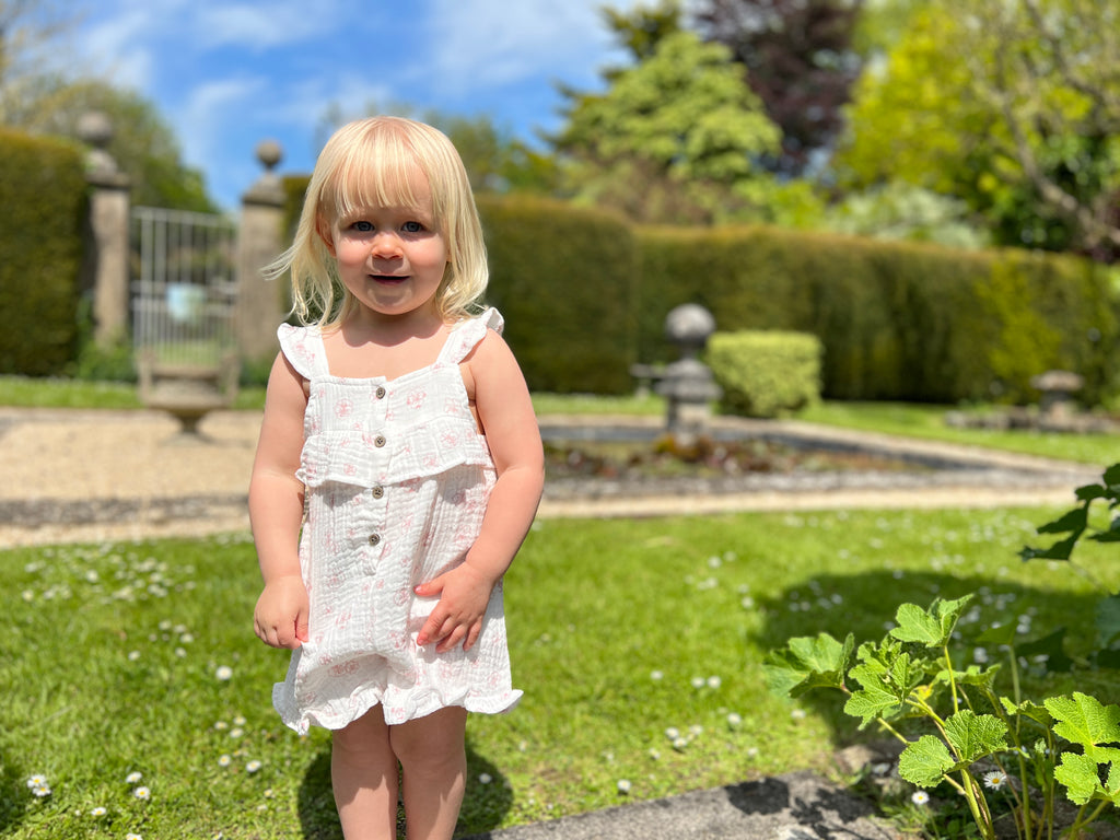 happy toddler standing in sunny garden wearing white gauze shortie overall with all over pink flower print. buttons on front and frill detail round front and legs