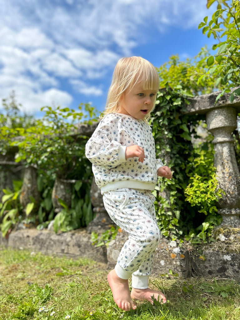 happy toddler walking in sunny garden in front of old stone wall wearing white sweatshirt with pastel flower print and matching pants