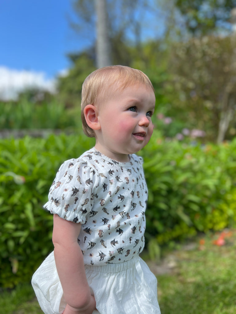 toddler standing in sunny garden wearing short sleeved white ribbed jersey onesie with seahorse print and white skort