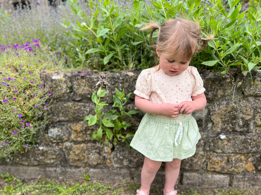 happy toddler in sunny garden standing in front of stone wall wearing delicate green skort and spotty cream top
