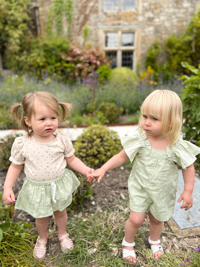 2 toddlers standing in front of garden stone wall holding hands one wearing happy toddler in sunny garden wearing delicate green skort and spotty cream top and the other delicate green short romper 