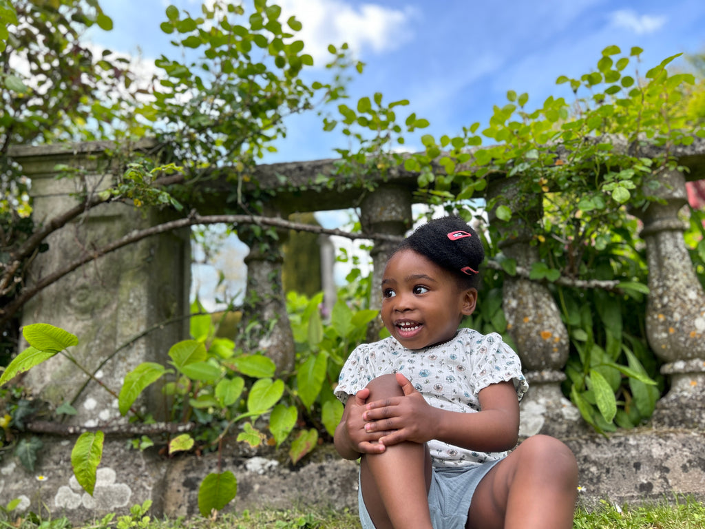 girl in garden sitting in front of old stone wall wearing tee shirt in white ribbed jersey with flower print and blue shorts