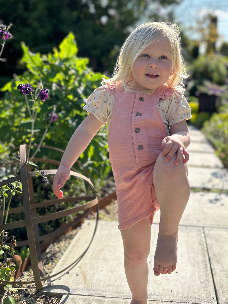 girl standing on path in garden on sunny day wearing pink shortie overalls with frill shoulder straps and buttons down the front with cream tee underneath