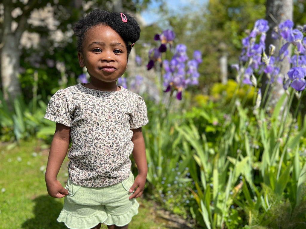 toddler standing in front of summer flowers on sunny day wearing tee shirt in cream ribbed jersey with bird and flower print and green shorts