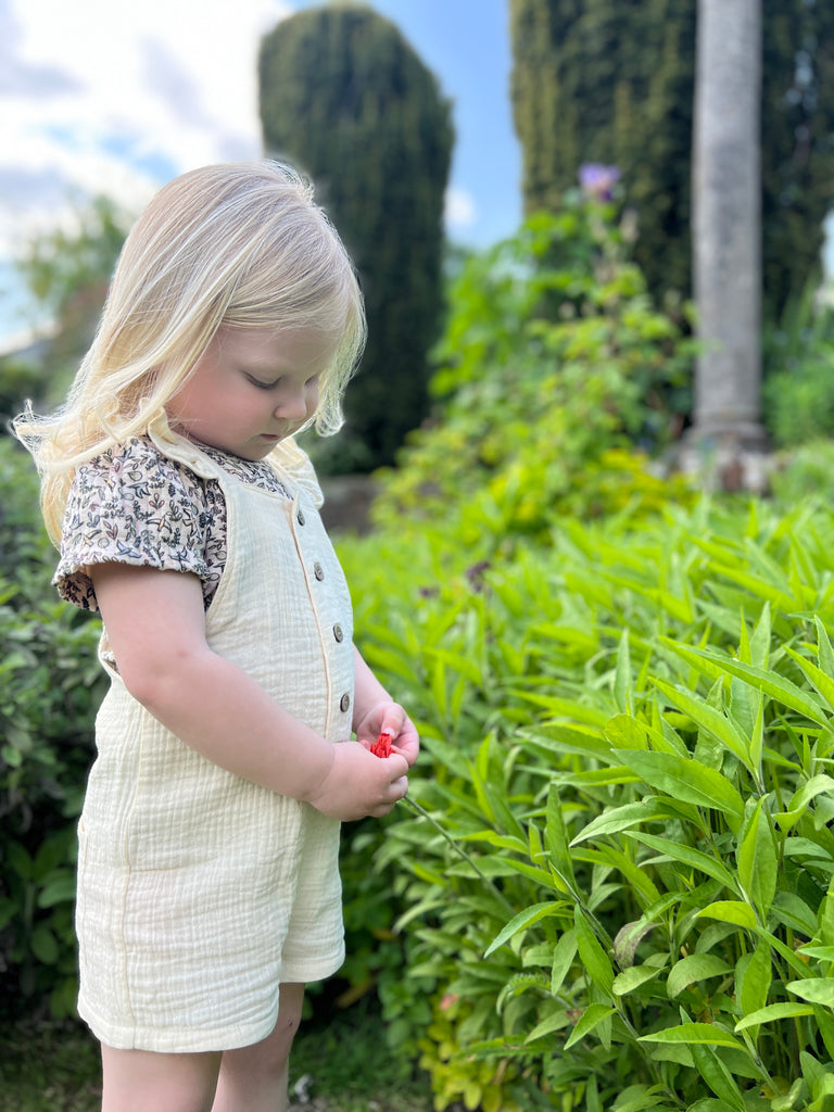girl standing in garden on sunny day wearing cream shortie overalls with frill shoulder straps and buttons down the front with print tee underneath