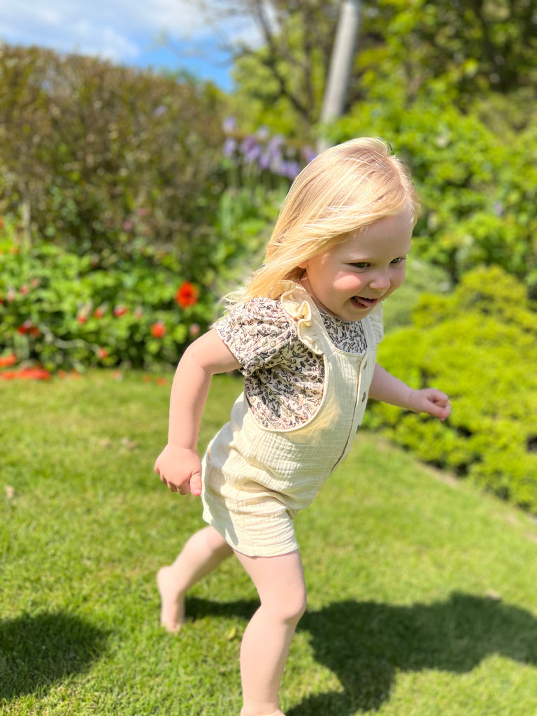 girl running in garden  on sunny day wearing cream shortie overalls with frill shoulder straps and buttons down the front with print tee underneath