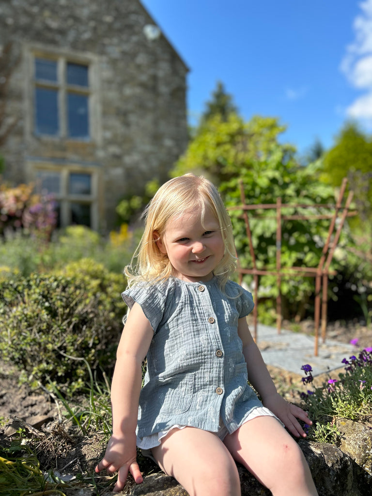 girl in sunny garden sat on wall wearing blue gauze blouse with capped sleeves buttons down the front 2 patch pockets on front seahorse print all over