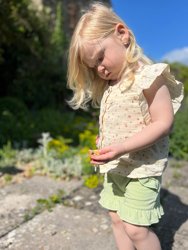 toddler standing on old stone path holding summer flowers on sunny day wearing frill sleeved blouse with square cut neck line and buttons down the front and green gauze shorts