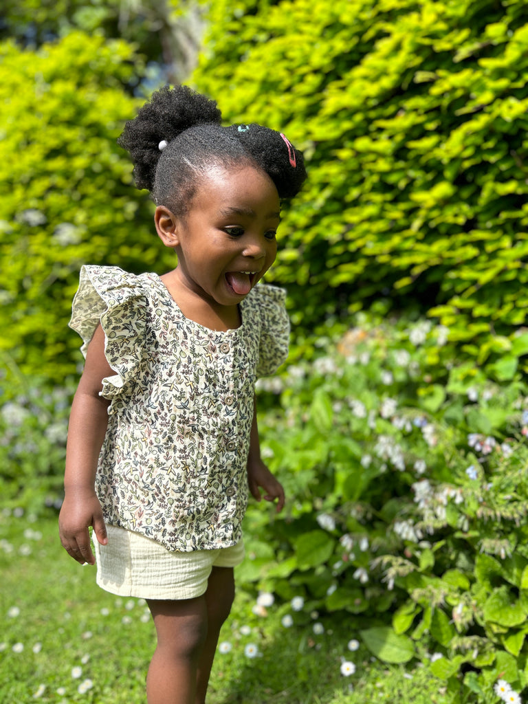 toddler standing on grass in front of large hedge on sunny day wearing frill sleeved blouse with flower and bird small print square cut neck line and buttons down the front