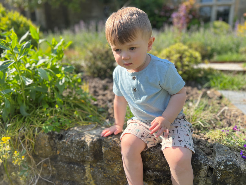 happy toddler in sunny garden wearing pale blue short sleeved henley tee with 2 button detail and cream shorts