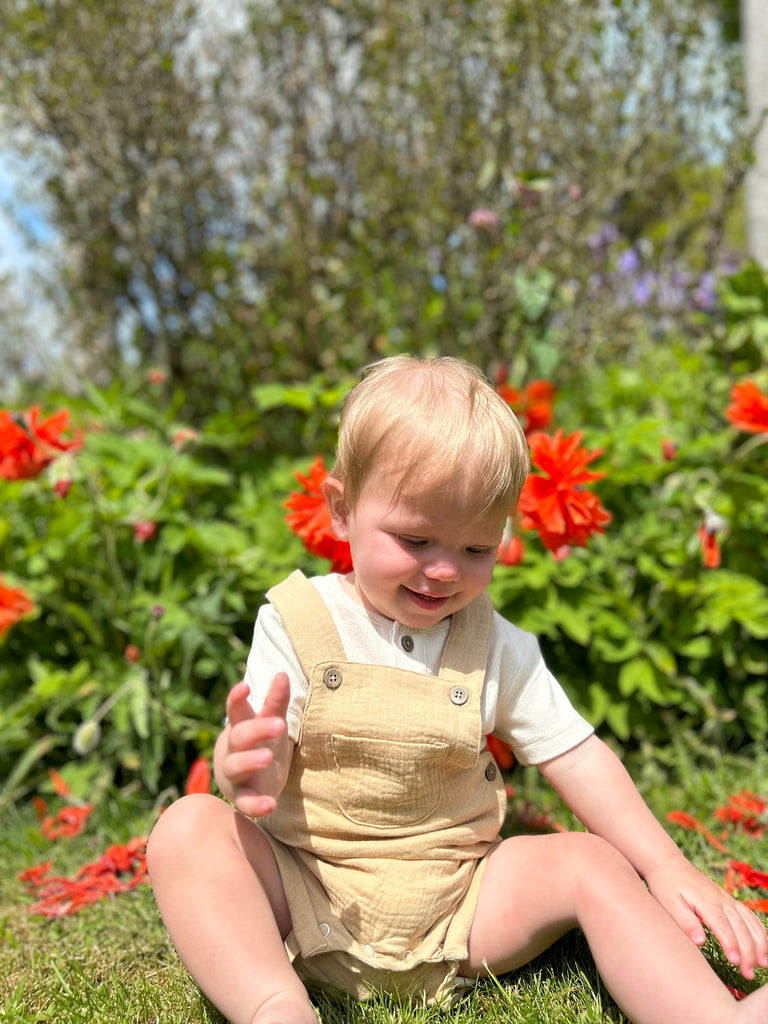 happy toddler sat in sunny garden in front of summer flowers wearing gold shortie gauze overalls