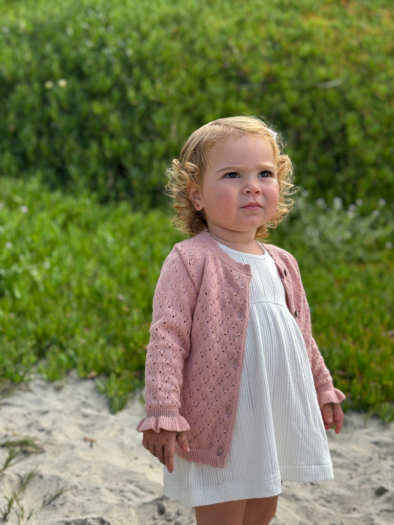 toddler standing on beach wearing white dress and pink knit cardigan with pretty pattern, scalloped edge around the neck and the bottom and cute frill cuffs with a gentle stretch wrist band. Buttons down the front.
