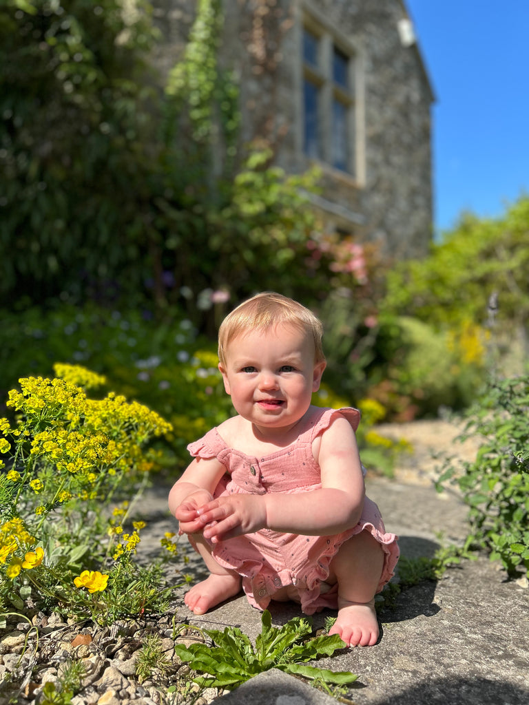 happy toddler in front of old stone manor house amongst summer flowers wearing  pale pink gauze shortie overal all over dotty print. buttons on front and frill detail round front and legs