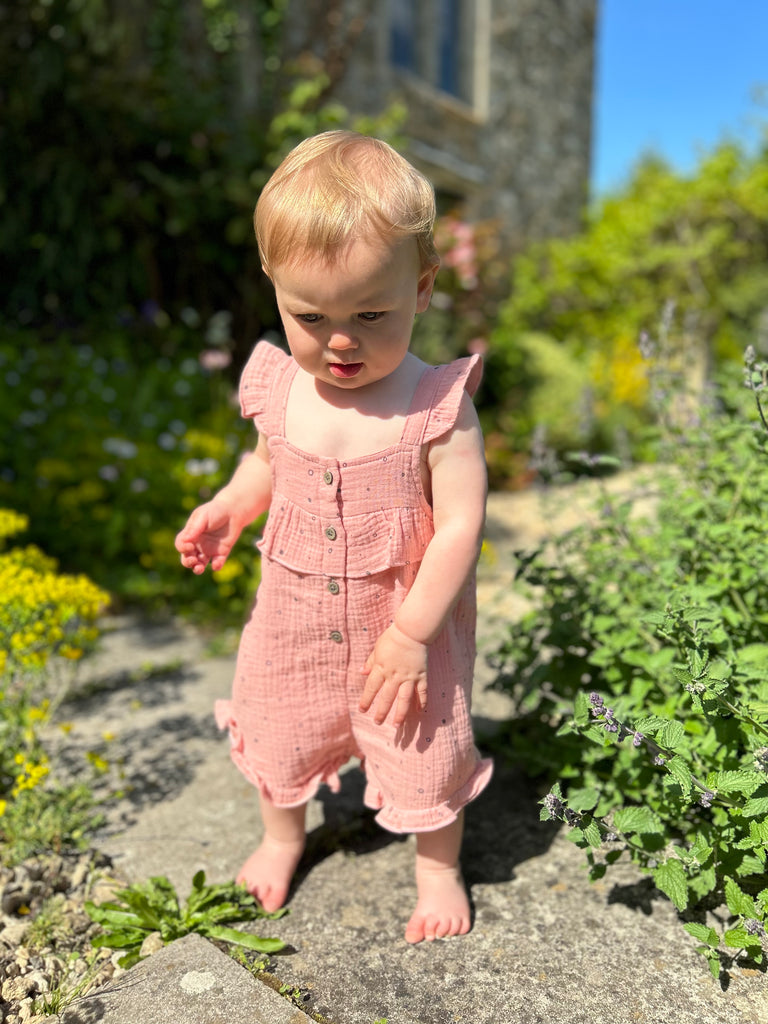 happy toddler standing in front of old stone manor house amongst flower beds wearing  pale pink gauze shortie overal all over dotty print. buttons on front and frill detail round front and legs