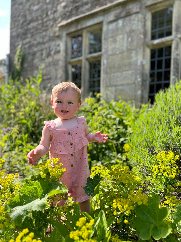 happy toddler standing in front of old stone manor house amongst flower beds wearing pale pink gauze shortie overal all over dotty print. buttons on front and frill detail round front and legs