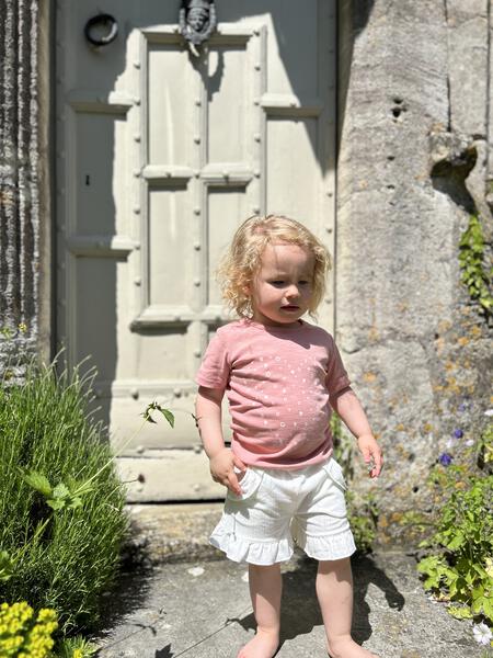 happy toddler standing in front of old wooden front door wearing pink short sleeve tee with heart dot print and white shorts