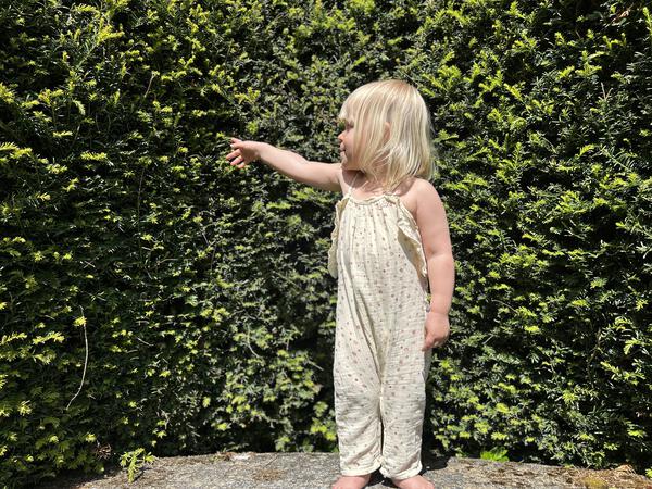 toddler standing infront of large hedge in sunny garden wearing jersey long legged romper with shoe string straps and frills pastel multi colored polka dots on cream background