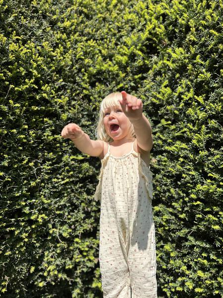 happy toddler standing infront of large hedge in sunny garden wearing jersey long legged romper with shoe string straps and frills pastel multi colored polka dots on cream background