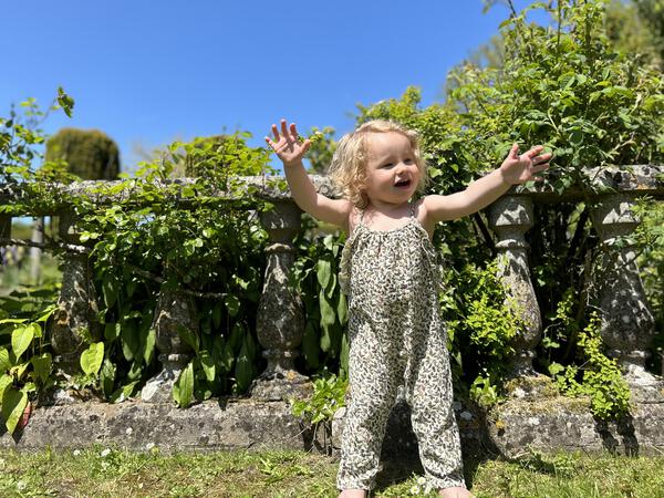 happy toddler standing in front of old stone wall  in sunny garden wearing  jersey long legged romper with shoe string straps and frills Small flowers and birds print on cream background