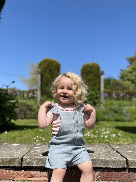 toddler in sunny garden sat on stone wall and wearing pink tee shirt with patterned fabric and frill neck under blue overalls