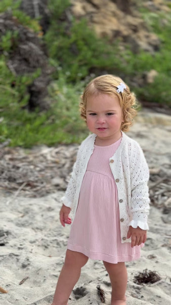 happy toddler on beach on sunny day wearing pale pink dress with frill over shoulders and short sleeves line pattern fabric and white knit cardigan