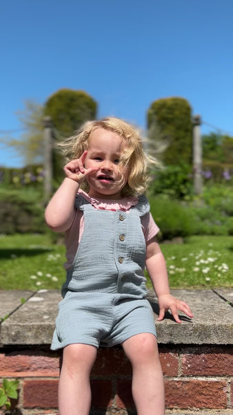 toddler in sunny garden sat on stone wall and wearing pink tee shirt with patterned fabric and frill neck under blue overalls