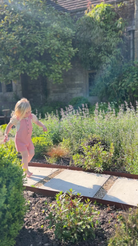 girl running down path in garden on sunny day wearing pink shortie overalls with frill shoulder straps and buttons down the front with cream tee underneath