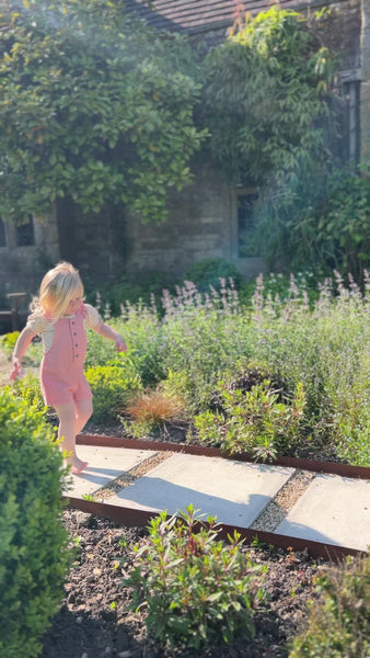 girl running down path in garden on sunny day wearing pink shortie overalls with frill shoulder straps and buttons down the front with cream tee underneath
