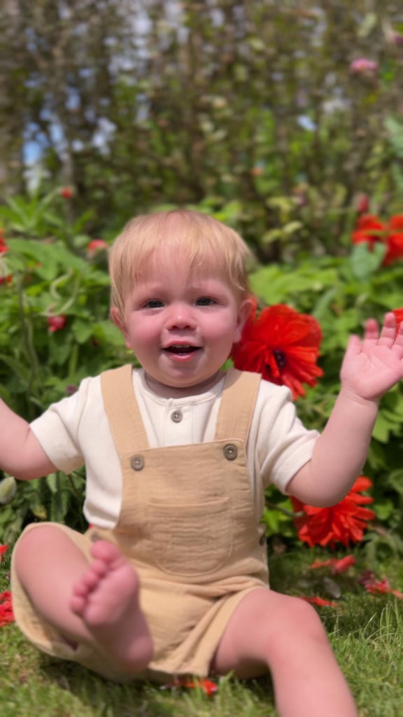 happy toddler sat in sunny garden in front of summer flowers wearing gold shortie gauze overalls
