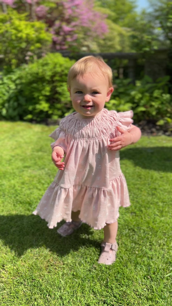 toddler walking on grass in sunshine on summer day wearing pale pink dress with white flower print smocked neck line short sleeves and large frill tier