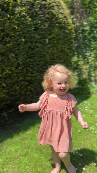 happy toddler laughing and running in garden on sunny day wearing coral dress in pointelle jersey with frill over shoulders