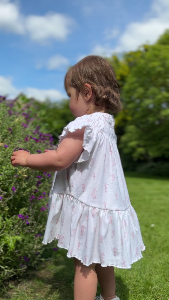 toddler standing in sunny garden in front of summer flowers wearing white dress with pink flower print smocked neck line short sleeves and large frill tier