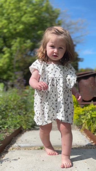 toddler in sunny garden standing amongst summer plants an d walking down garden path wearing white dress in cotton jersey with frill over shoulders in guitar print