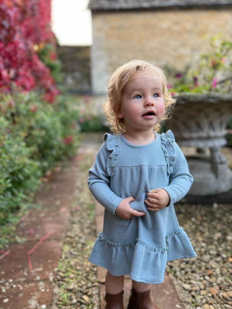 toddler girl standing on path wearing blue ribbed long sleeve dress with ruffles over the shoulders and tiered frill on bottom of dress