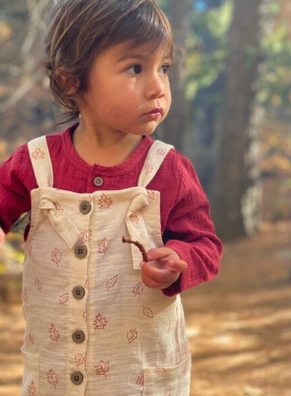 toddler standing in sunny autumn woods wearing cream overalls with leaf all over print. knotted straps and buttons down the front bib. patch pockets at side and red gauze top underneath