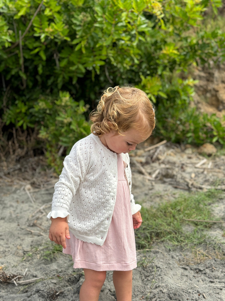 toddler standing on beach wearing pink dress and white knit cardigan with pretty pattern, scalloped edge around the neck and the bottom and cute frill cuffs with a gentle stretch wrist band. Buttons down the front.
