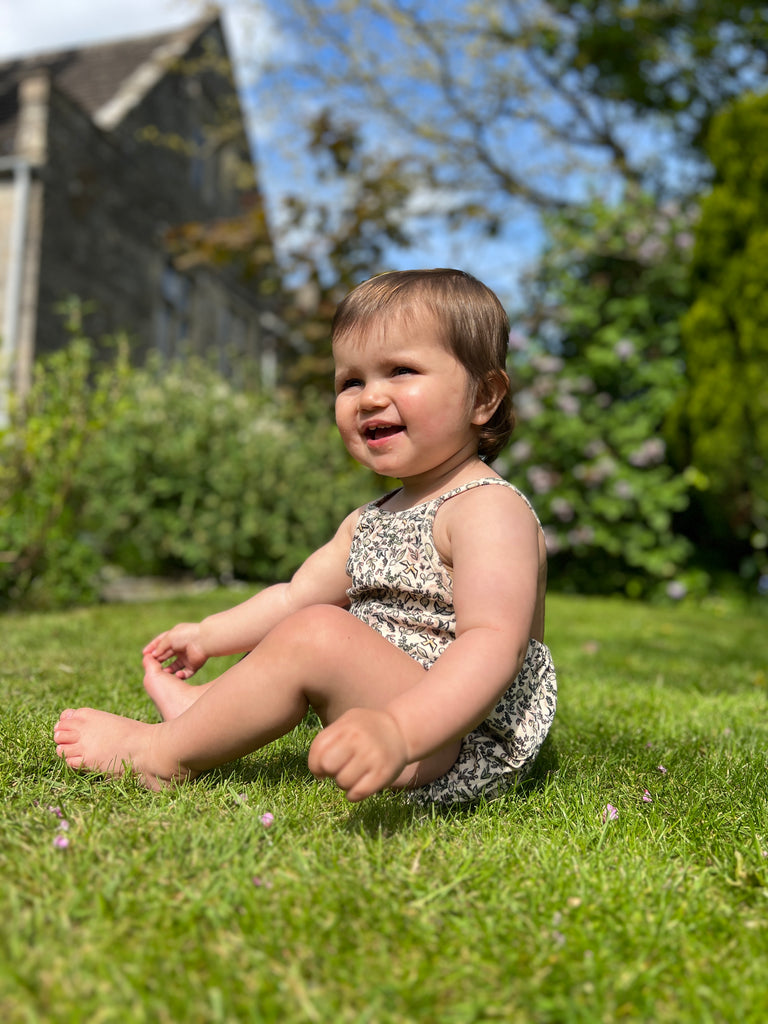 happy baby sat on grass on sunny day wearing cream bubble with shoe string straps in flower and bird print