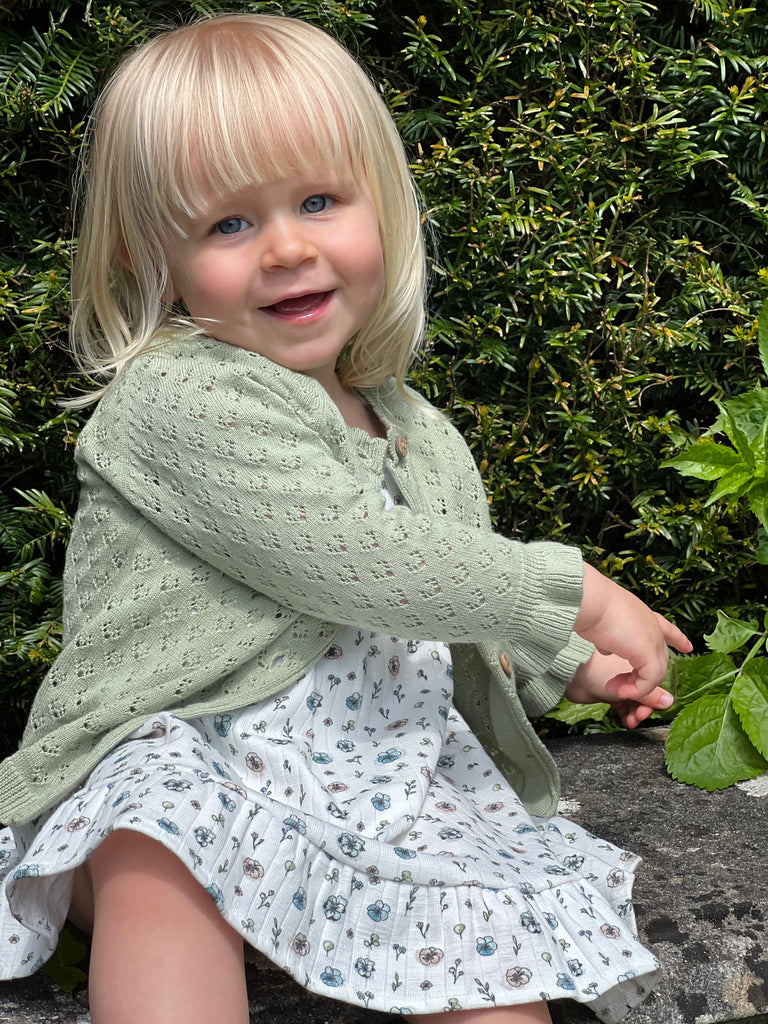 happy toddler sat in front of bush wearing sage green lace knit cardigan frill cuffs on sleeve and flower dress