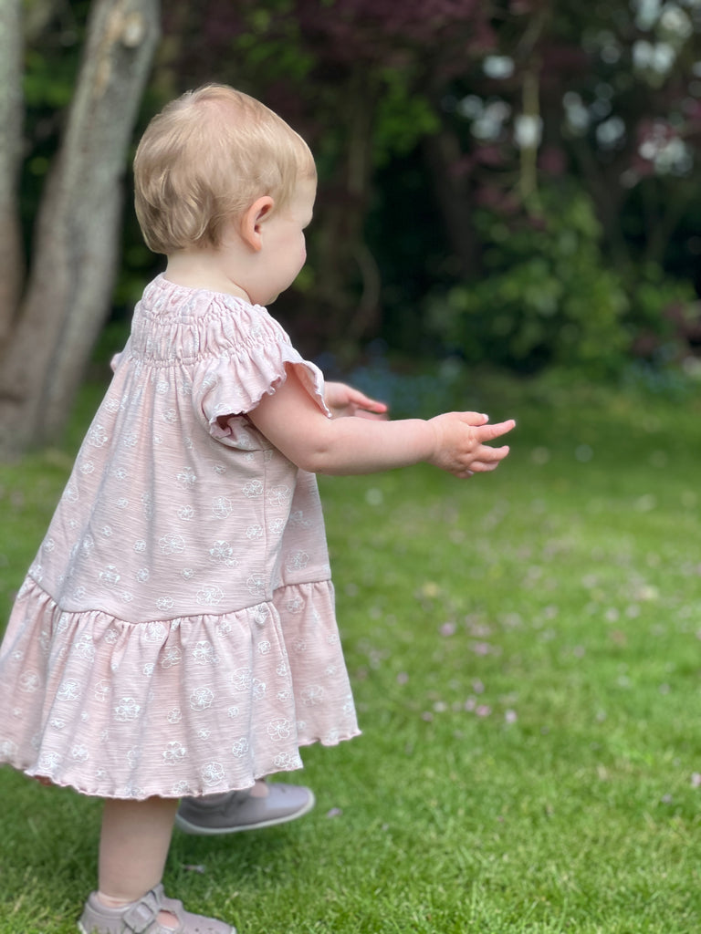 toddler standing on grass on summer day wearing pale pink dress with white flower print smocked neck line short sleeves and large frill tier