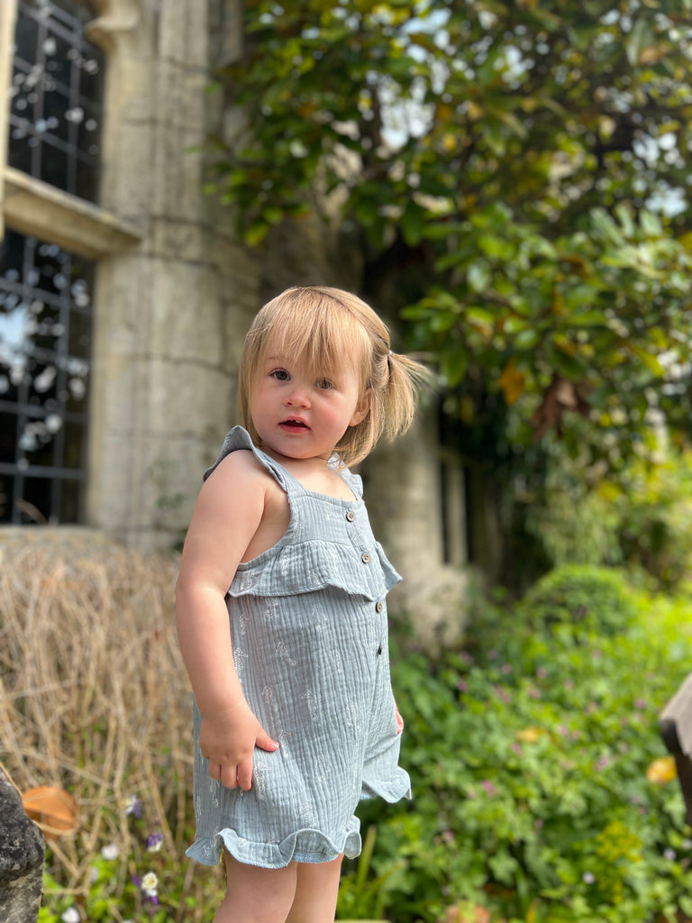 toddler standing in front of old stone window of manor house wearing gentle blue gauze shortie overalls with white all over seahorse print. buttons on front and frill detail round front and legs