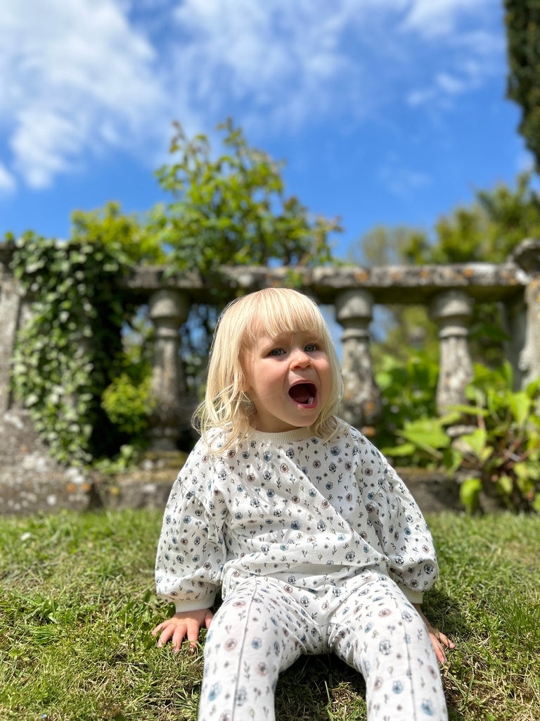 happy toddler sat in sunny garden in front of old stone wall wearing white sweatshirt with pastel flower print and matching pants