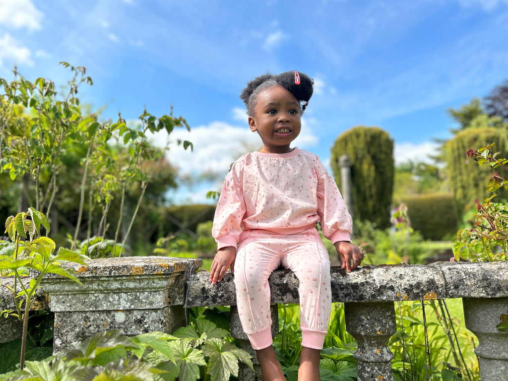 happy toddler sat on old stone wall in sunny garden wearing pink sweatshirt with pastel polka dots print and matching pants