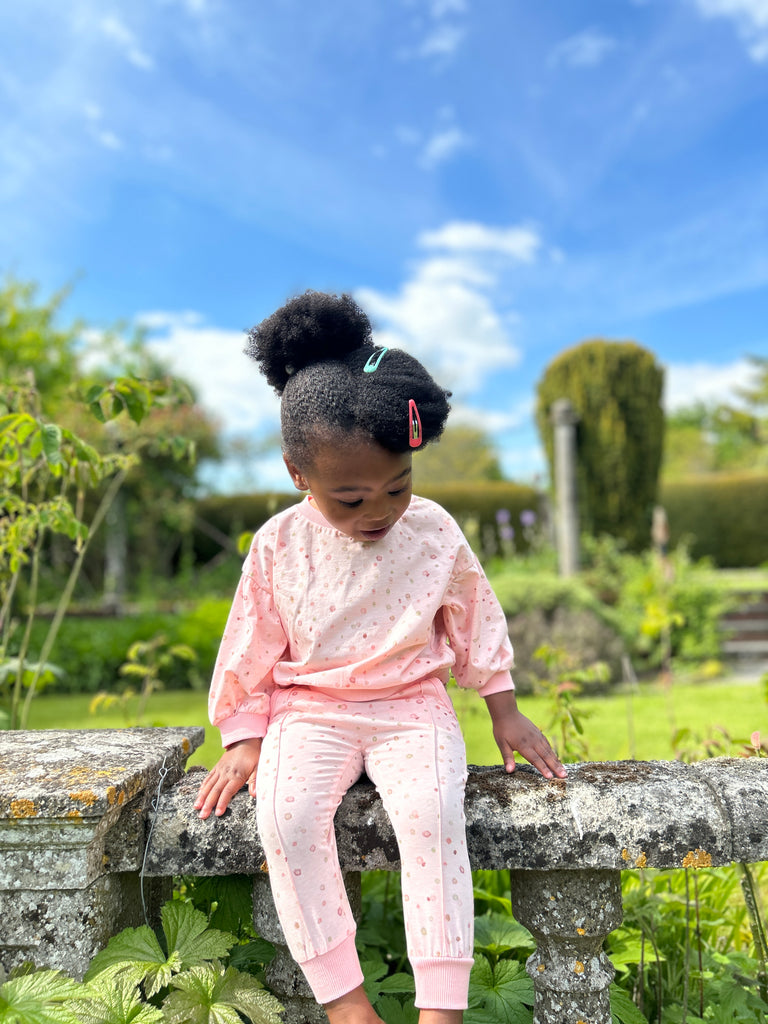 toddler sat on old stone wall on sunny day wearing pink polka dot joggeres with elastic waistband and drawcord side pockets and front seam detail and matching top