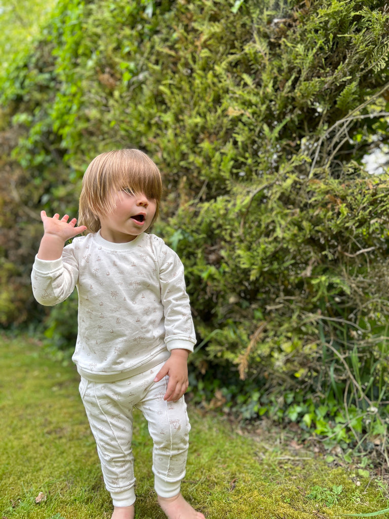toddler walking on grass on sunny day wearing white palm tree and island print joggers with elastic waistband and drawcord side pockets and front seam detail and matching top