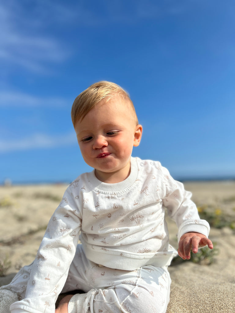 toddler on beach on sunny day with big blue sky wearing white sweatshirt with all over cream island and boat print poppers at neck and matching pants