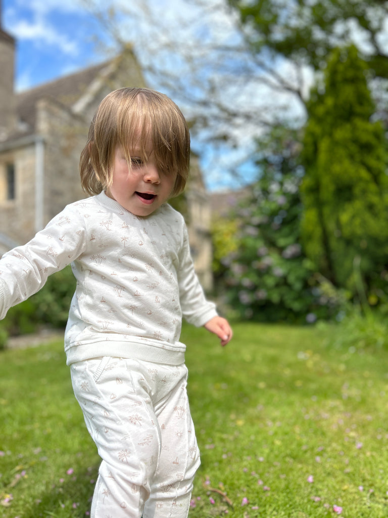 toddler walking on grass on sunny day wearing white palm tree and island print joggers with elastic waistband and drawcord side pockets and front seam detail and matching top