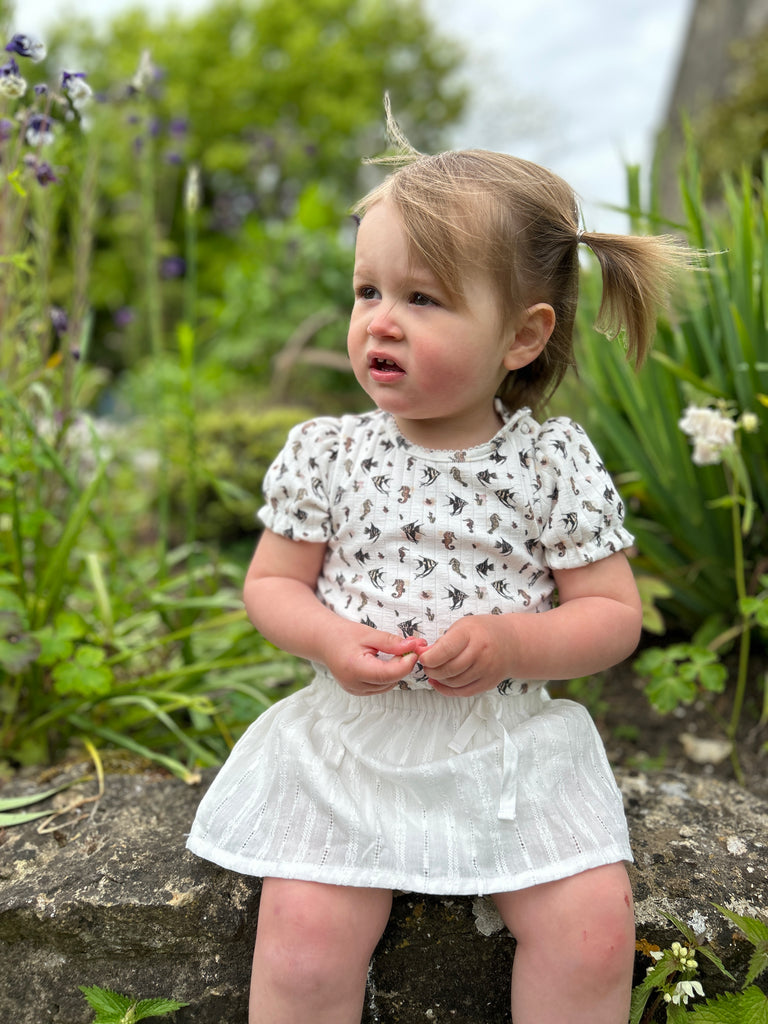 happy toddler sat on stone garden wall wearing delicate white skort and white seahorse print top
