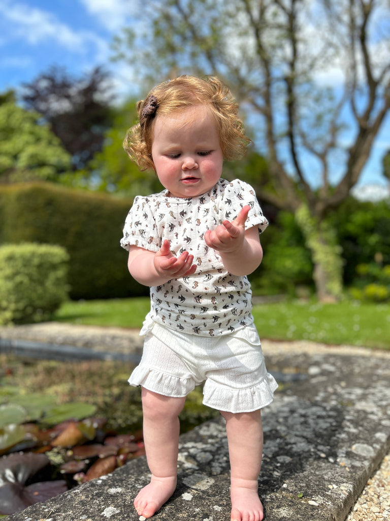 toddler in sunny garden wearing white gauze shorts with 2 side pockets and frill around legs and white tee